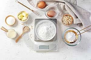 Flour in white bowl measuring on digital scale with baking ingredients and utensil on marble kitchen table