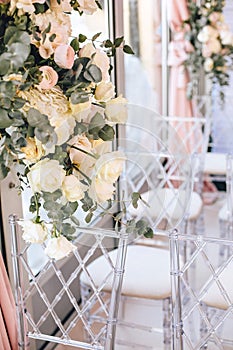 Flower arrangement of white roses and eucalyptus next to transparent chairs adorns the wedding ceremony zone in classical style