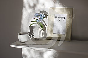 Flower hackelia velutina in white fluted vase in the style of the seventies, a photo frame with the words Enjoy, and a cup of