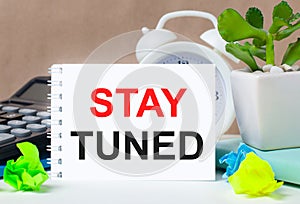 Flower in a pot, calculator, white alarm clock, multi-colored pieces of paper and a white notebook with the text STAY TUNED on the
