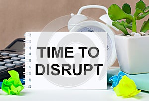 Flower in a pot, calculator, white alarm clock, multi-colored pieces of paper and a white notebook with the text TIME TO DISRUPT