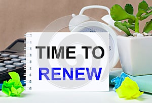 Flower in a pot, calculator, white alarm clock, multi-colored pieces of paper and a white notebook with the text TIME TO RENEW on