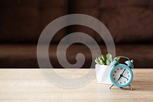 Flower pot and clock on wooden table against defocused sofa with pillows. Front view. Good morning concept. Mock-up