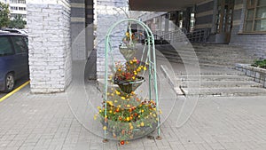 Flower pots are suspended on a metal frame set on a tile platform. Nearby, cars are parked on the roadway and there is an entrance