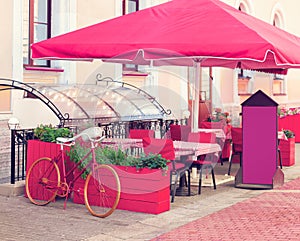 A flowerbed made from a bicycle, and tubs of flowers in a summer cafÃ©. Stone sidewalk, tables, chairs, protective awning. Photo.