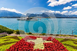 Flowerbed of the Swiss flag with boat cruise on the Thun lake and Alps mountains, Oberhofen, Switzerland. Swiss flag made of