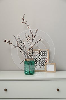 Flowering tree twigs in glass vase with decor on white chest of drawers near light wall