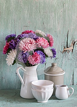 Flowers asters in a white enameled pitcher and vintage crockery - ceramic bowl and enameled jar, on a blue wooden background.