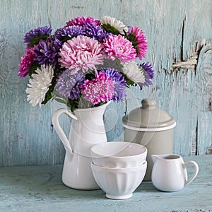 Flowers asters in a white enameled pitcher and vintage crockery - ceramic bowl and enameled jar, on a blue wooden background.