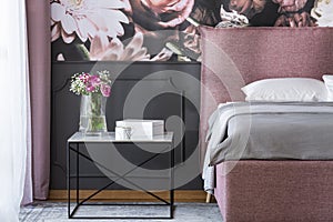 Flowers on black table next to pink and grey bed in bedroom interior with wallpaper. Real photo
