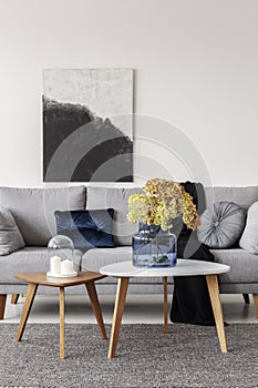 Flowers in blue glass vase and two white candles on wooden coffee tables in grey elegant living room with comfortable sofa