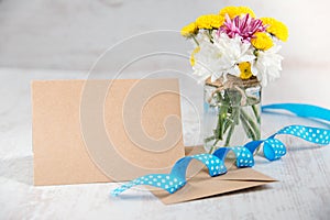 Flowers bouquet in a jar vase with card note, envelope and blue ribbon on a white wood rustic background