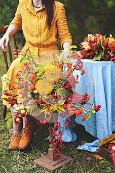 Flowers bouquet of orange, red, yellow dahlias. Brunette woman in yellow dress, maple leaves wreath on wooden chair by round table