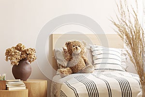 Flowers in brown vase on wooden nightstand table next to single bed with stripped bedding with teddy bear