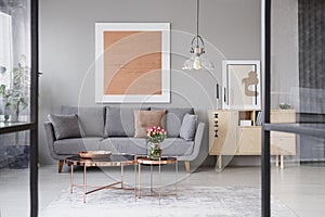 Flowers on copper table in front of grey couch in living room interior with rose gold poster. Real photo