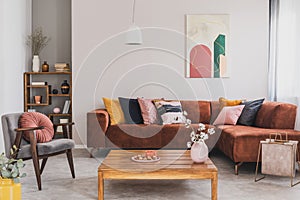 Flowers in vase on wooden coffee table in fashionable living room interior with brown corner sofa with pillows and abstract photo