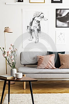 Flowers on wooden table in front of grey sofa with pillows in flat interior with posters. Real photo photo