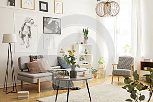 Flowers on wooden table next to grey couch in living room interior with lamp and posters. Real photo