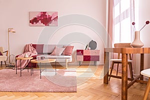 Flowers on wooden table in pink apartment interior with sofa under poster next to cabinet. Real photo