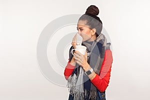 Flu treatment. Portrait of unhealthy young woman wrapped in warm scarf cleaning nose with napkin and holding tea cup