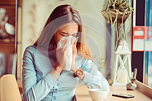 Flu. Young woman got nose allergy, flu sneezing nose sitting at the table in a trendy cafe coffee shop with a cup of hot beverage