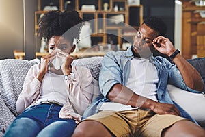 Flus no fun at all. a young woman blowing her nose with her husband sitting next to her on the sofa.