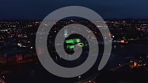 Fly above night city, green illuminated large building on Shannon river waterfront. Limerick, Ireland
