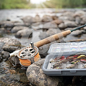 A fly fishing rod and an open fly fishing box lie on the rocks of a mountain river at sunset.