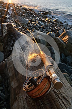 A fly fishing rod and an open fly fishing box lie on the sea rocks at sunset.