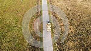 We fly over a tractor with weaves of hay driving along the road into the field