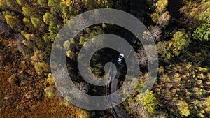 Flying above a white car driving on colorful autumn forest road. Aerial drone view