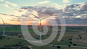 Flying behind a working wind turbine with the sun in front of it amidst a cloudy sky at sunset in a rural setting with green