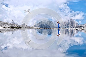 Flying bird and a woman in blue dress walking along the alpine lake with beautiful reflections of mountains and clouds