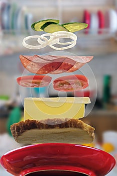 Flying layers of sandwich with ham, cheese, vegetables over the red plate with blurred kitchen background. Breakfast food concept