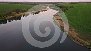 Flying over the smooth water of the river with reflection of pink sunset among green fields