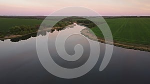 Flying over the smooth water of river with the reflection of a pink sunset among green fields