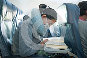 Flying in times of covid19 - young sweet cute Asian Chinese woman in face mask sitting on airplane cabin reading book or novel