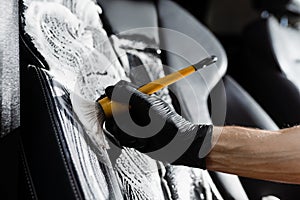 Foam cleaning black leather seat using brush. Worker in auto cleaning service clean car inside. Car interior detailing.
