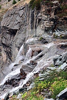 Foaming cascades of an alpine waterfall on granite rocks of various textures and colors overgrown with sparse green vegetation