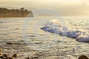 Foaming wave with backwash breaking across bay with rocky shoreline, penninsulla, sandy beach