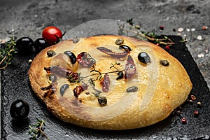 Foccacia, Italian bread, with olive oil, olives, sun-dried tomatoes, capers and thyme, Mediterranean bread on a dark background.