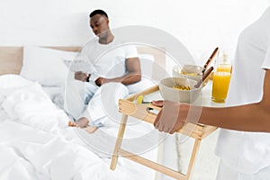 Focus of african american woman holding tray with breakfast, standing near bed in bright room