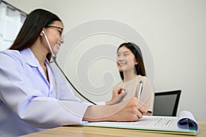 Focus of Asian female doctor hand checking heart rate of young female patient with stethoscope and taking note on prescription of