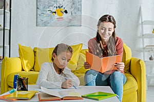 Focus of attractive babysitter holding notebook near cute child studying at home