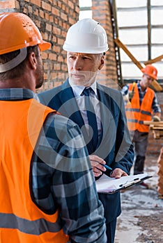 Focus of bearded businessman in helmet holding clipboard near constructors