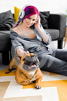 Focus of beautiful girl with colorful hair and headphones sitting on floor and petting french bulldog