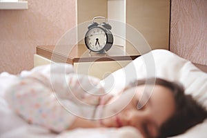 Focus on black alarm clock showing half past six, on bedside nightstand, in blurred foreground of a sleeping child girl
