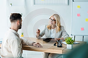 Focus of blonde recruiter looking at man and holding clipboard with pen