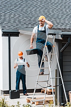 Focus of builder touching helmet and standing with toolbox on ladder near coworker