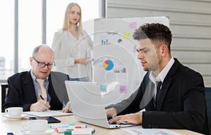 Focus on Business manager wearing eyeglass writing on paper and staff sitting typing and seeing on laptop near colour pens. Woman